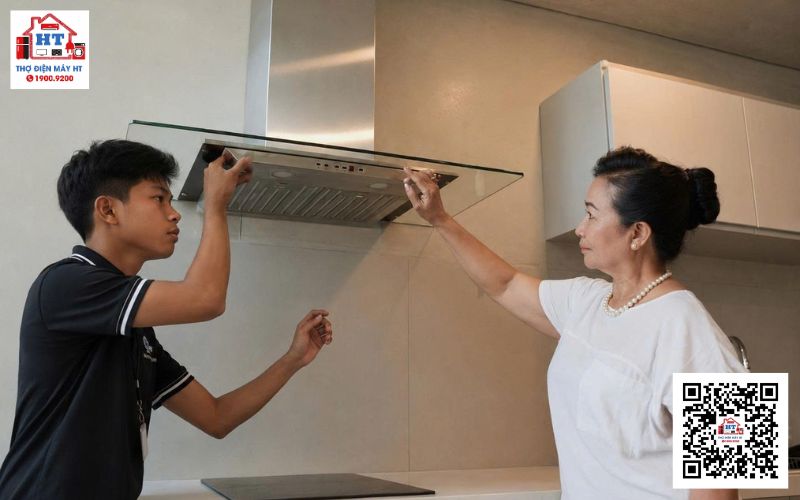 Kiểm tra tổng quát máy hút mùi Người thợ đang kiểm tra máy hút mùi gắn tường. Anh ấy mặc áo xanh, tay cầm dụng cụ chuyên dụng.